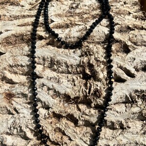 Puede incluir: Un collar largo de cuentas negras con un dise&ntilde;o delicado. El collar est&aacute; compuesto por peque&ntilde;as cuentas facetadas, creando un efecto brillante. Las cuentas est&aacute;n ensartadas, formando un bucle continuo. El collar se muestra sobre una superficie marr&oacute;n texturizada.