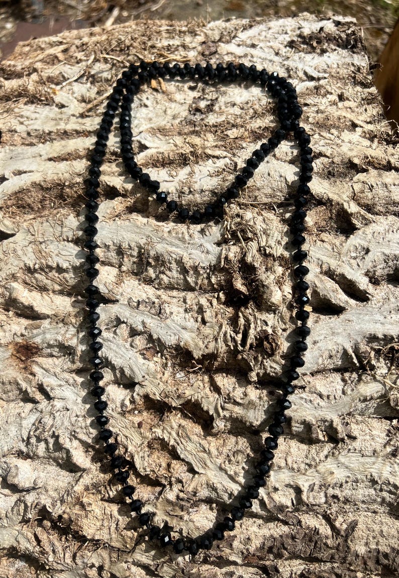 Puede incluir: Un collar largo de cuentas negras con un dise&ntilde;o delicado. El collar est&aacute; compuesto por peque&ntilde;as cuentas facetadas, creando un efecto brillante. Las cuentas est&aacute;n ensartadas, formando un bucle continuo. El collar se muestra sobre una superficie marr&oacute;n texturizada.