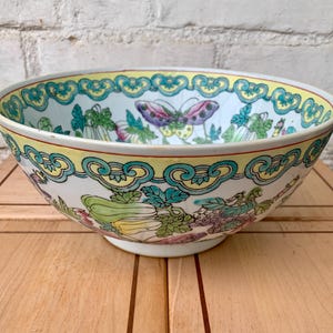 Puede incluir: Un cuenco decorativo de cerámica con base blanca y borde ancho. El cuenco presenta un colorido diseño floral y de mariposas con detalles en amarillo, verde, morado y azul. El cuenco está sobre una superficie de madera.
