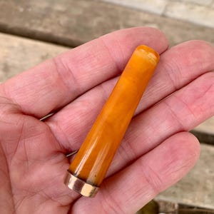 May include: A cylindrical, amber-colored object with a polished surface and a gold-colored metal band at the base. The object is held in a person's hand, and the background is a wooden surface.