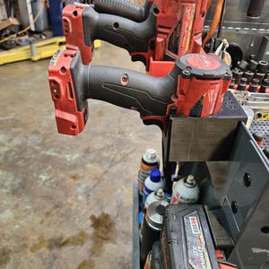 May include: Two red and black cordless impact wrenches are mounted on a tool cart. The tools have black grips and are labeled "FUEL". A black battery pack is visible below the tools, along with various spray cans.