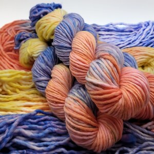 May include: Close-up of colorful yarn skeins in shades of peach, yellow, and blue. The yarn is thick and textured, with a soft, inviting appearance. The arrangement suggests a craft or knitting project.