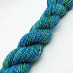 Op de afbeelding: Een gedraaide streng garen in tinten van teal, blauw en groen. Het garen lijkt een zachte, natuurlijke vezel te zijn, met subtiele kleurvariaties over de hele streng. Het garen is strak gedraaid, wat suggereert dat het geschikt is voor brei- of haakprojecten.