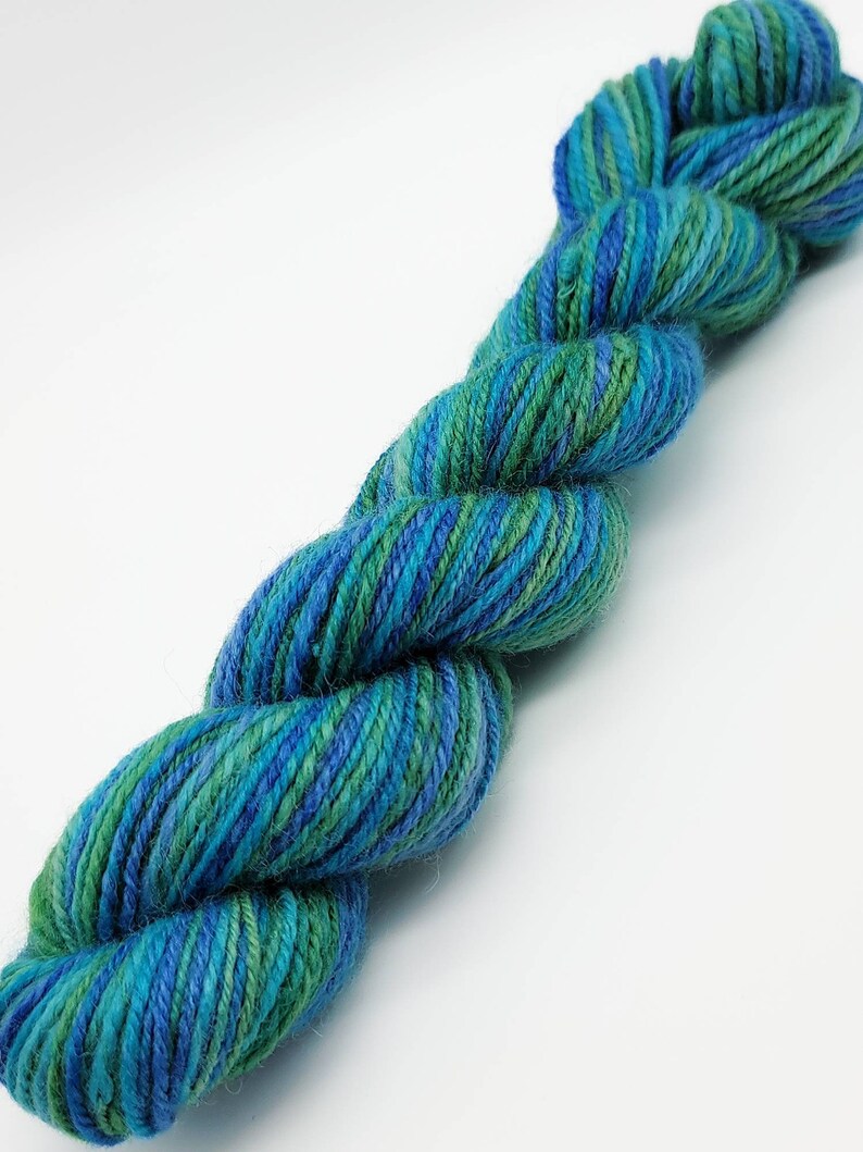 Op de afbeelding: Een gedraaide streng garen in tinten van teal, blauw en groen. Het garen lijkt een zachte, natuurlijke vezel te zijn, met subtiele kleurvariaties over de hele streng. Het garen is strak gedraaid, wat suggereert dat het geschikt is voor brei- of haakprojecten.