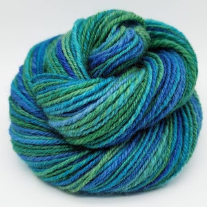 Op de afbeelding: Een bol garen in tinten blauw en groen. Het garen lijkt een mix van kleuren te zijn, met delen van diepblauw, lichtblauw en verschillende tinten groen. Het garen is strak opgerold, wat suggereert dat het klaar is om te breien of te haken.