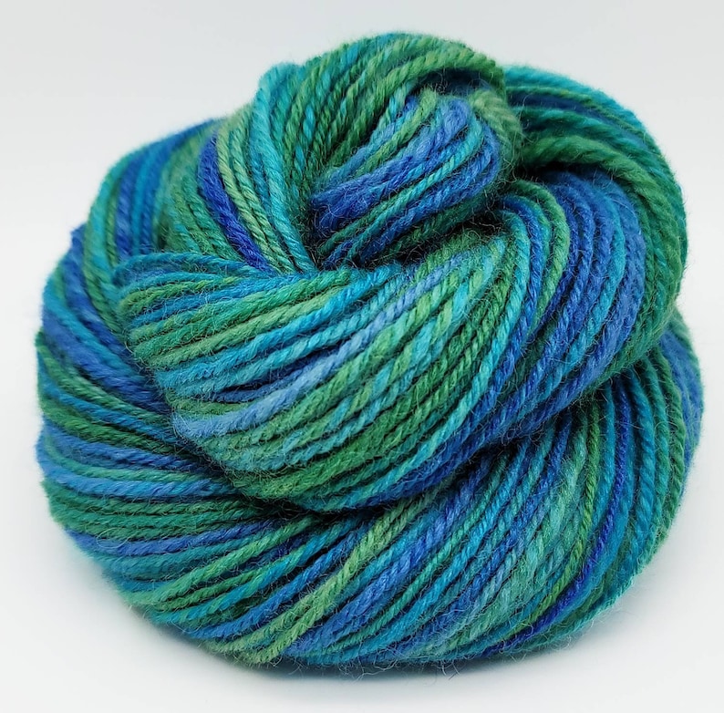 Op de afbeelding: Een bol garen in tinten blauw en groen. Het garen lijkt een mix van kleuren te zijn, met delen van diepblauw, lichtblauw en verschillende tinten groen. Het garen is strak opgerold, wat suggereert dat het klaar is om te breien of te haken.