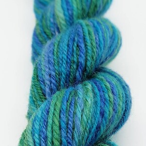 Op de afbeelding: Close-up van een streng garen in tinten blauw en groen. Het garen is strak opgewonden en toont een mix van kleuren. Het garen wordt waarschijnlijk gebruikt voor brei- of haakprojecten.