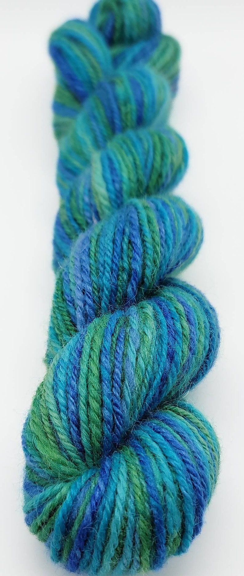 Op de afbeelding: Close-up van een streng garen in tinten blauw en groen. Het garen is strak opgewonden en toont een mix van kleuren. Het garen wordt waarschijnlijk gebruikt voor brei- of haakprojecten.