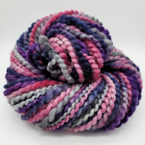 May include: A ball of colorful yarn in shades of pink, purple, blue, and gray. The thick yarn is tightly wound, creating a textured appearance. Ideal for knitting or crocheting projects.