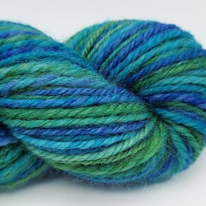 Op de afbeelding: Een close-up van een bol garen in tinten blauw en groen. Het garen is strak opgerold, met zichtbare strengen en een textuur. De kleuren vloeien in elkaar over, waardoor een visueel aantrekkelijke en natuurlijke uitstraling ontstaat, geschikt voor brei- of haakprojecten.