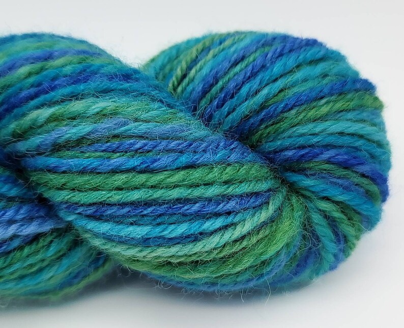 Op de afbeelding: Een close-up van een bol garen in tinten blauw en groen. Het garen is strak opgerold, met zichtbare strengen en een textuur. De kleuren vloeien in elkaar over, waardoor een visueel aantrekkelijke en natuurlijke uitstraling ontstaat, geschikt voor brei- of haakprojecten.