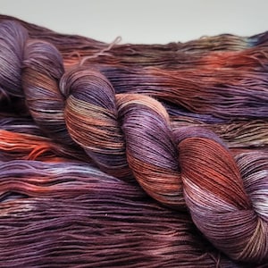 May include: A skein of hand-dyed yarn in shades of purple, orange, and brown. The yarn is twisted into a skein, with loose strands visible in the background. The colors blend together, creating a variegated effect, suitable for knitting or crochet projects.