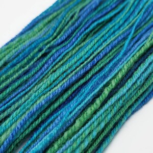 Op de afbeelding: Close-up van kleurrijk garen in tinten blauw en groen. De garenstrengen zijn strak gedraaid, waardoor een textuur ontstaat. De kleur overgangen cre&euml;ren een gradi&euml;nteffect, waarbij donkerdere blauw- en groentinten in elkaar overlopen. Ideaal voor brei- of haakprojecten.