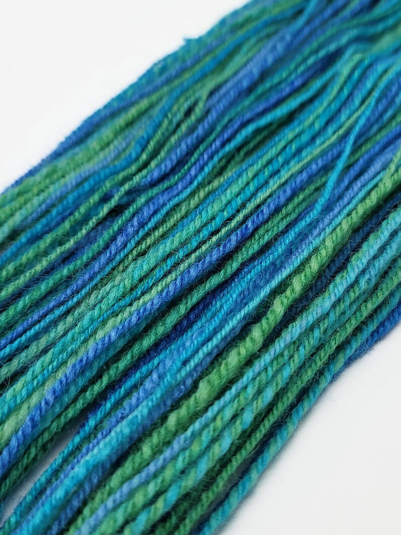 Op de afbeelding: Close-up van kleurrijk garen in tinten blauw en groen. De garenstrengen zijn strak gedraaid, waardoor een textuur ontstaat. De kleur overgangen cre&euml;ren een gradi&euml;nteffect, waarbij donkerdere blauw- en groentinten in elkaar overlopen. Ideaal voor brei- of haakprojecten.