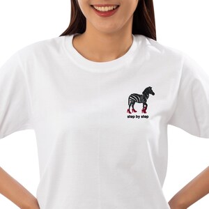 Könnte beinhalten: Weißes T-Shirt mit einem schwarz-weißen Zebra, das rote Schuhe trägt und den Text "step by step" auf der Vorderseite gestickt hat.