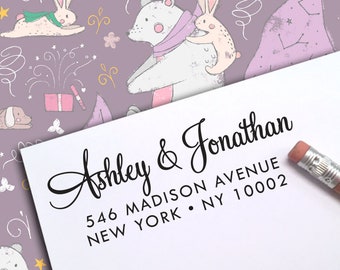 May include: A white card with the names "Ashley & Jonathan" in elegant black script, along with an address. The background features a whimsical pattern of cartoon animals, stars, and swirls in soft pastel colors. A pencil rests on the card.