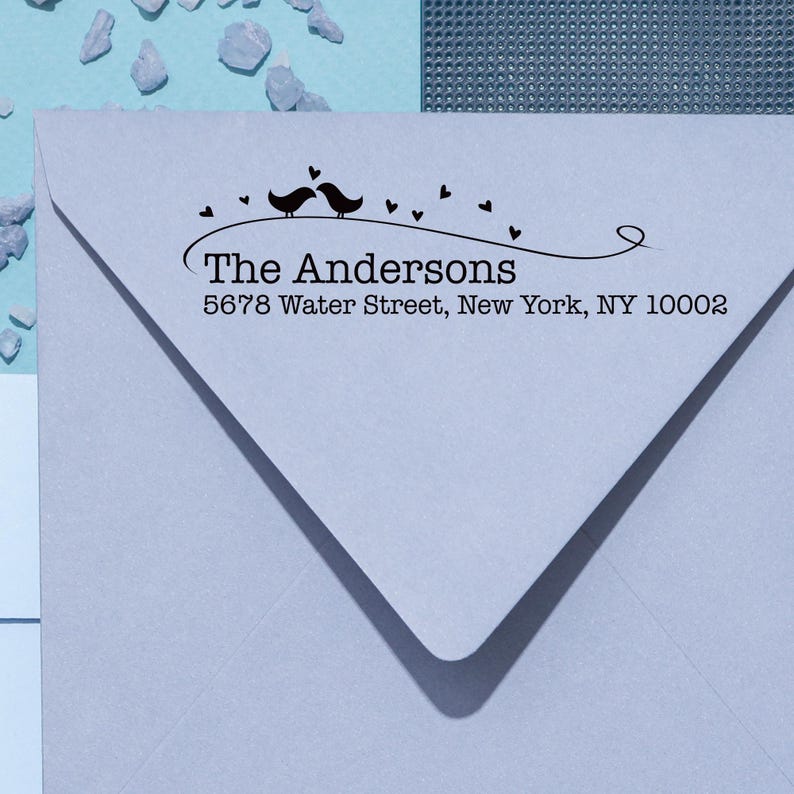 Peut inclure: Une enveloppe bleu clair avec un motif noir repr&eacute;sentant deux oiseaux, des c&oelig;urs et le texte "The Andersons" avec une adresse. Le motif est centr&eacute; sur l'enveloppe.