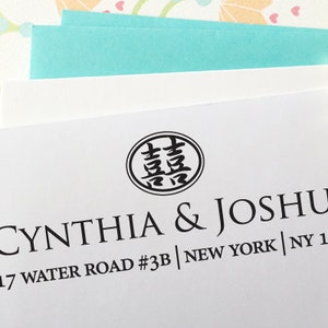 May include: White invitation with black text reading "CYNTHIA & JOSHUA" and address details. A circular emblem with Chinese characters is above the names. Gold-colored geometric beads are in the background, along with pastel stationery.