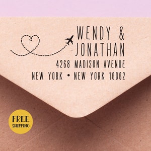 Puede incluir: Un sobre marrón con un sello de goma negro que dice "WENDY & JONATHAN 4268 MADISON AVENUE NEW YORK • NEW YORK 10002" con un pequeño avión y un corazón conectados por una línea punteada. Un círculo amarillo con el texto "ENVÍO GRATIS" está en la esquina inferior izquierda.