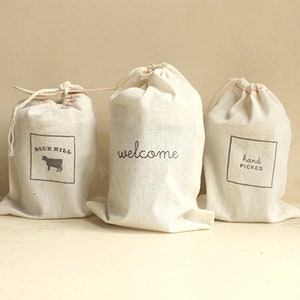 May include: Three white fabric drawstring bags with different text printed on them. The first bag says "Blue Hill" with a silhouette of a cow. The second bag says "welcome". The third bag says "hand picked".