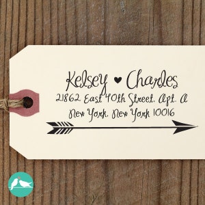 以下が含まれることがあります： 黒い文字で「Kelsey ♥ Charles」と住所詳細が書かれたクリーム色のタグ。黒い矢印が右を指しています。タグにはバーガンディ色の補強があり、ひもで結ばれています。左下隅には、ターコイズと白のロゴがあります。