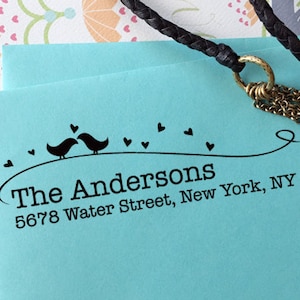 Peut inclure: Une enveloppe bleu clair avec un tampon d'adresse noir. Le tampon repr&eacute;sente deux oiseaux, des c&oelig;urs et le texte "The Andersons, 5678 Water Street, New York, NY 10002". Un cordon tress&eacute; noir et une cha&icirc;ne dor&eacute;e sont dans l'image.
