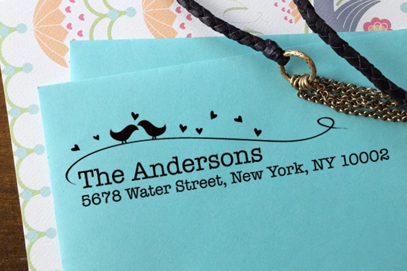 Peut inclure: Une enveloppe bleu clair avec un tampon d'adresse noir. Le tampon repr&eacute;sente deux oiseaux, des c&oelig;urs et le texte "The Andersons, 5678 Water Street, New York, NY 10002". Un cordon tress&eacute; noir et une cha&icirc;ne dor&eacute;e sont dans l'image.