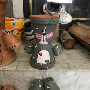 Peut inclure: Une figurine décorative fabriquée à partir de pots en terre cuite gris empilés, peinte pour ressembler à un chat. Le motif présente un ventre blanc avec un petit cœur rouge, des joues roses et des taches blanches. Bras et jambes articulés.