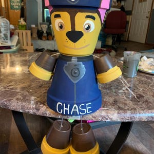 Op de afbeelding: Een handgeschilderde terracotta plantenpot in de vorm van het personage Chase uit de cartoon Paw Patrol. De pot is geschilderd in tinten geel, bruin, blauw en zwart. De pot heeft een blauwe hoed met een roze rand en een witte voetafdruk. De pot is gelabeld met "CHASE".
