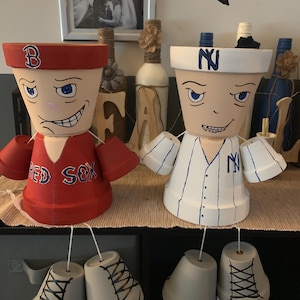 May include: Two painted terracotta flower pots are decorated to look like baseball players. The pot on the left is painted red with the words "Red Sox" and the letter "B" on the hat. The pot on the right is painted white with the letter "N" on the hat and the words "NY" on the jersey. Both pots have painted legs and shoes.