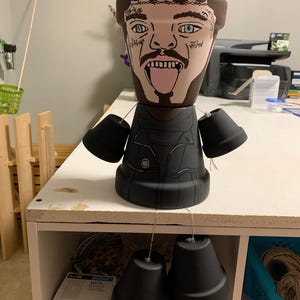 Puede incluir: Un macetero de cerámica marrón con un retrato pintado de una persona con tatuajes y la lengua fuera. El macetero está decorado con pintura negra para parecer una chaqueta. Cuatro maceteros de cerámica negros más pequeños cuelgan del macetero principal.