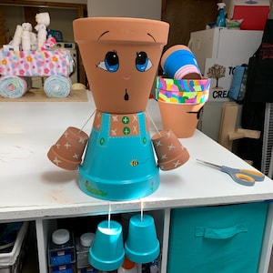Puede incluir: Una figura de maceta de terracota pintada a mano. La figura tiene una cabeza marrón con ojos azules y una expresión de sorpresa. Lleva un peto turquesa con una abeja y detalles verdes. Dos macetas más pequeñas sirven como brazos y dos macetas turquesas cuelgan como piernas.