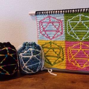 Op de afbeelding: Vier gehaakte dobbelstenen zakken in verschillende kleuren, waaronder zwart, blauw, groen en roze, met een gehaakt wandkleed met vier dobbelstenen in dezelfde kleuren.