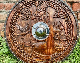 May include: A round wooden shield with a leather trim and metal studs. The shield is decorated with carvings of a bird, a dragon, a ship, and a warrior holding a spear. The shield also has a metal center piece and the text "STAM FOR PNIFTS RVNIR".