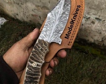 May include: A hunting knife with a brown and black stag horn handle and a leather sheath. The blade is made of Damascus steel with a wavy pattern. The sheath has the text "ModelKnives" printed on it.