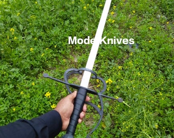 May include: A black and white sword with a decorative guard and a black handle. The sword is held by a hand in a field of green grass and yellow flowers. The text "Mode Knives" is visible in the image.