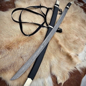 May include: A black leather scabbard with a silver-coloured metal hilt and a long, curved, silver-coloured blade with a damascus pattern. The sword is lying on a brown and white fur surface.