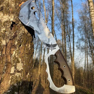 May include: A silver and black tactical axe with a brown handle. The axe has a serrated edge and a hole in the blade. The axe is leaning against a tree trunk. The text "MODEL KNIVES" is engraved on the blade.