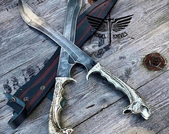 May include: Two crossed, silver-bladed daggers with ornate gold-colored handles rest on a weathered wooden surface. A leather sheath with red and black accents is visible. The text "MODEL KNIVES" is printed on the sheath.