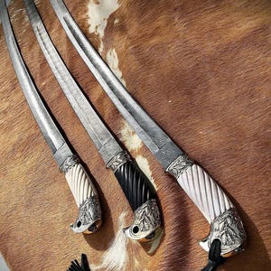 K&ouml;nnte beinhalten: Drei verzierte Schwerter mit silbernen Klingen und kunstvollen Griffen. Die Griffe sind aus wei&szlig;en, schwarzen und braunen Materialien gefertigt. Die Schwerter werden auf einer braunen und wei&szlig;en Pelzfl&auml;che pr&auml;sentiert.