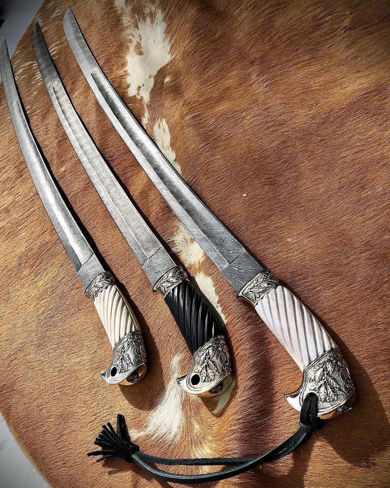 K&ouml;nnte beinhalten: Drei verzierte Schwerter mit silbernen Klingen und kunstvollen Griffen. Die Griffe sind aus wei&szlig;en, schwarzen und braunen Materialien gefertigt. Die Schwerter werden auf einer braunen und wei&szlig;en Pelzfl&auml;che pr&auml;sentiert.