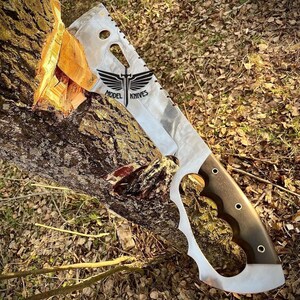 May include: A large, silver, serrated knife with a black handle and a wing design with the text "MODEL KNIVES". The knife is partially inserted into a tree trunk.