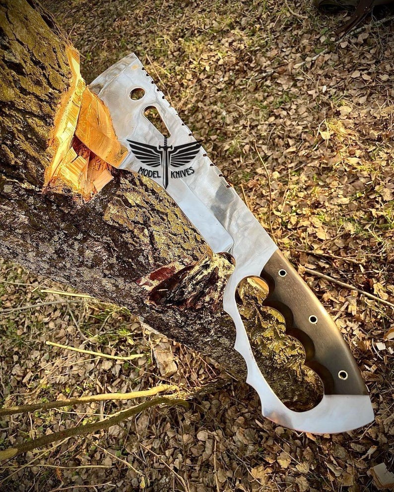May include: A large, silver, serrated knife with a black handle and a wing design with the text "MODEL KNIVES". The knife is partially inserted into a tree trunk.
