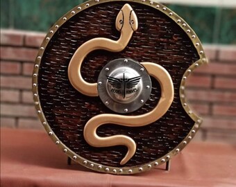May include: A round, wooden shield with a dark brown finish and a gold-colored snake design. The shield has a metal rim and a small, silver emblem in the center with the text "MODEL KNIVES".