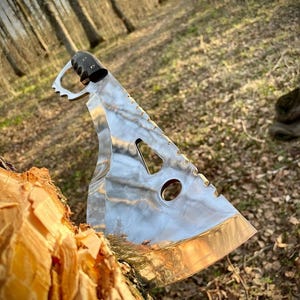 May include: A silver machete with a black handle and a serrated edge. The machete is partially inserted into a tree trunk.