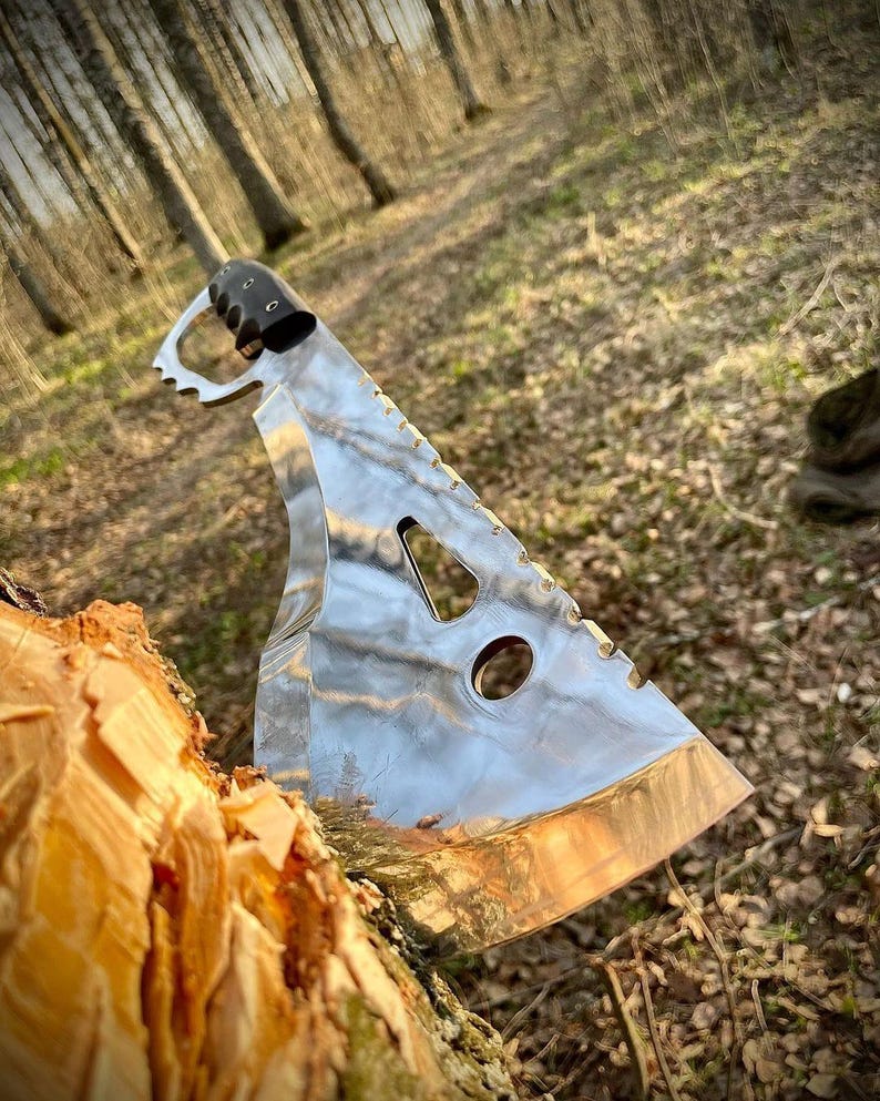 May include: A silver machete with a black handle and a serrated edge. The machete is partially inserted into a tree trunk.