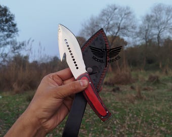 May include: A stainless steel knife with a red handle and a black leather sheath. The knife blade has a unique shape with a series of holes and a hook-like tip. The sheath is embossed with a logo that reads "MODEL KNIVES".