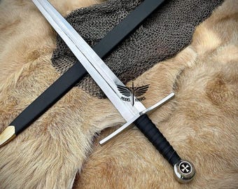 May include: A silver sword with a black leather grip and a cross pommel. The sword is lying on a brown fur surface with a black leather scabbard and a silver helmet in the background.