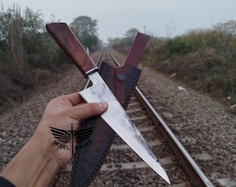 May include: A handcrafted chef's knife with a dark wooden handle and a stainless steel blade. The knife is paired with a brown leather sheath. The knife is held over a set of railroad tracks.