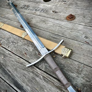 Könnte beinhalten: Ein mittelalterliches Schwert mit polierter Stahlklinge und einem braunen, lederumwickelten Griff. Das Schwert liegt auf einer Holzoberfläche neben einer beigefarbenen Scheide mit goldfarbenen Akzenten. Der Parier ist silberfarben.